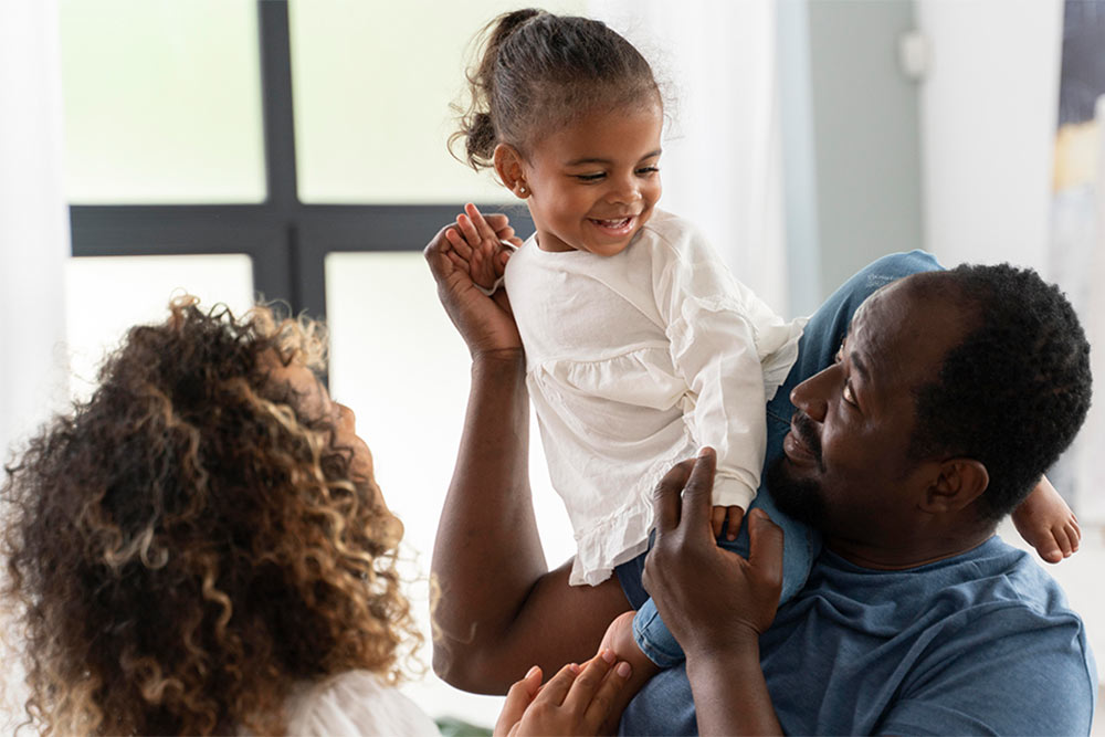 man holding his young daughter on his shoulder, with the mother facing the child, smiling