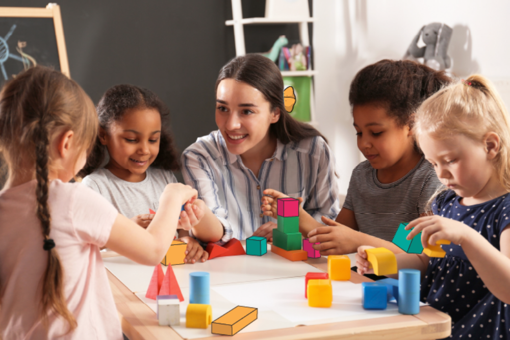 Adult facilitator and five young children sit around a table, collaboratively stacking and arranging colourful wooden blocks and geometric shapes in a classroom setting, with shelves and toys visible in the background.