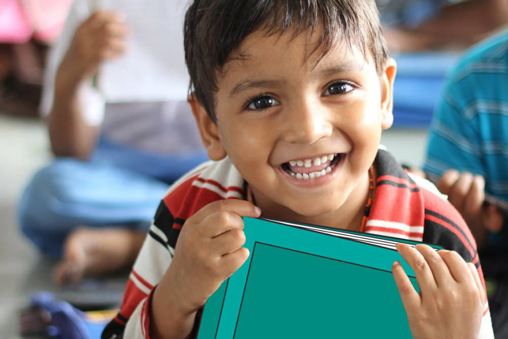 Close-up of a young child holding a teal-colored book toward the camera, seated indoors with other children and classroom materials visible out of focus in the background.