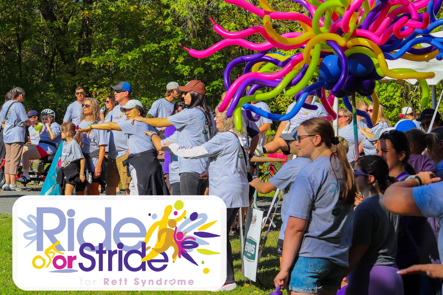 Ride for Stride participants gather outdoors beneath colourful balloon decorations during a community event for Rett Syndrome.